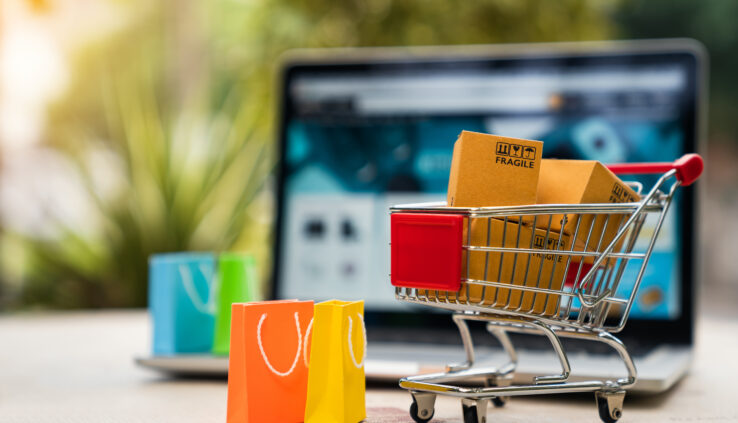 Product package boxes in cart with shopping bag and laptop computer with blurred web store shop on screen for online shopping and delivery concept