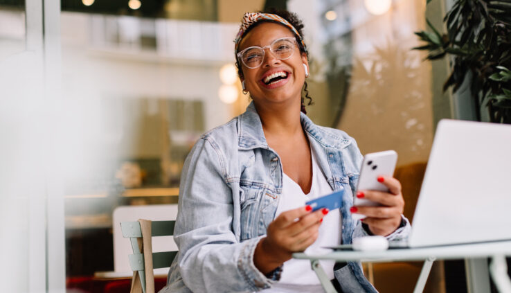 Young woman in a café happily uses her smartphone for online shopping, paying with a credit card.