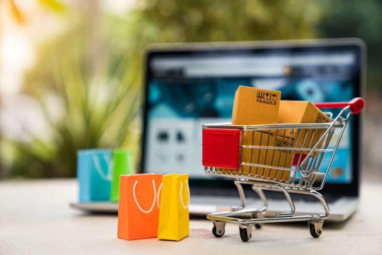 Product package boxes in cart with shopping bag and laptop computer with blurred web store shop on screen for online shopping and delivery concept