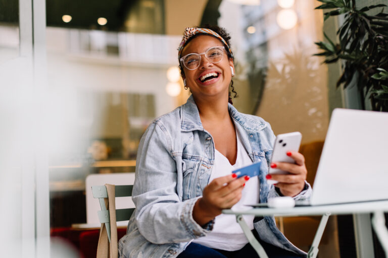 Young woman in a café happily uses her smartphone for online shopping, paying with a credit card.